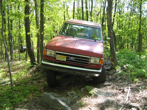 1990 Ford Bronco - Red Ford Bronco off-roading on a rocky trail through a wooded area.