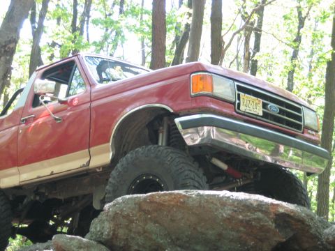 1990 Ford Bronco - A red Ford Bronco navigates rocky terrain in a wooded area, its lifted suspension clearly visible.