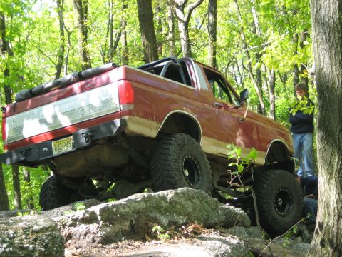 1990 Ford Bronco - A modified Ford Bronco tackles a rocky obstacle on an off-road trail, with onlookers observing from the side.