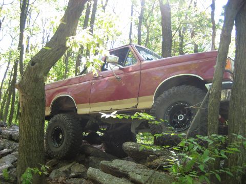 1990 Ford Bronco - A red 1990 Ford Bronco tackles a challenging rocky terrain in a wooded area.