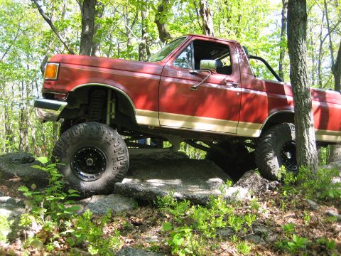 1990 Ford Bronco - This modified SUV is shown off-roading on a rocky trail surrounded by trees.