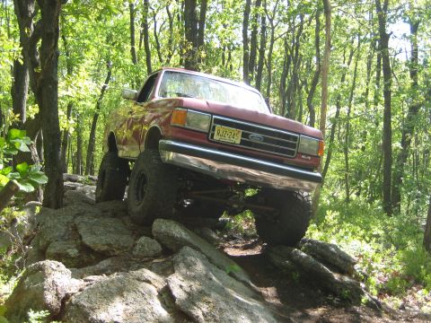 1990 Ford Bronco - Red 1990 Ford Bronco navigating a rocky off-road course in the woods.