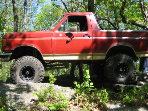 1990 Ford Bronco - This red Ford Bronco is navigating a rocky trail with its suspension lifted and large off-road tires.