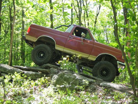 1990 Ford Bronco - A red 1990 Ford Bronco navigates a rocky trail through a dense forest.