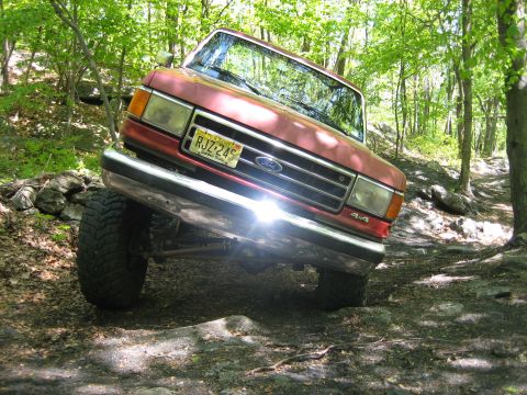 1990 Ford Bronco - Red 1990 Ford Bronco navigating a challenging off-road trail with trees and rocks.
