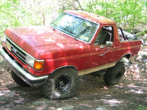 1990 Ford Bronco - This red Ford Bronco is shown off-roading on a dirt and rock path surrounded by trees.