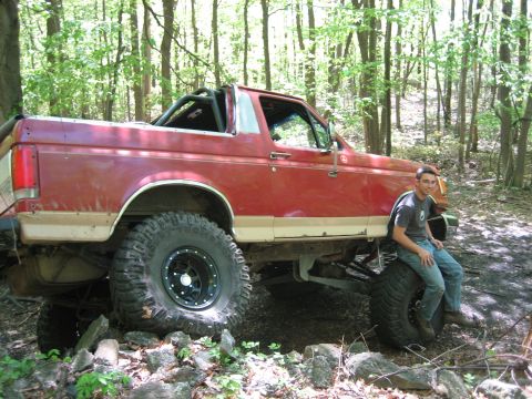 1990 Ford Bronco - Modified red Ford Bronco with off-road tires sits in a wooded area, with a man resting on the front wheel.
