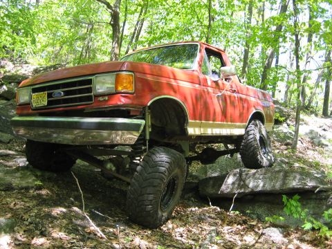 1990 Ford Bronco - Red 1990 Ford Bronco navigating a rocky trail in a wooded area.