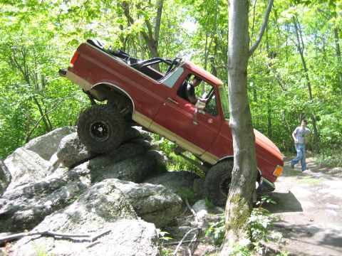 1990 Ford Bronco - This red SUV is attempting to climb over large rocks on a wooded trail.