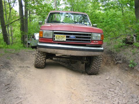1990 Ford Bronco - This 1990 Ford Bronco is captured from the front, navigating a wooded dirt path with its 4x4 designation visible.