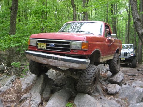 1990 Ford Bronco - A red Ford pickup truck navigates rocky terrain off-road, with a Jeep following behind.
