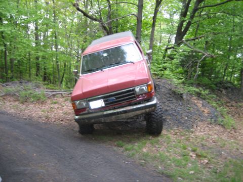 1990 Ford Bronco - A red Ford truck navigates an off-road trail, tilted precariously on a muddy incline.
