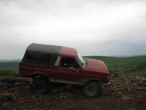 1990 Ford Bronco - A lifted Ford Bronco with a soft top navigates an off-road trail with a scenic, hilly landscape in the background.