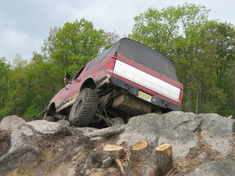 1990 Ford Bronco - This red 1990 Ford Bronco is shown tackling a challenging off-road course with large rocks and trees in the background.