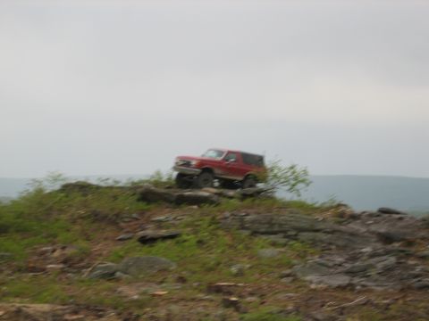1990 Ford Bronco - A red SUV navigates a rocky incline under an overcast sky.
