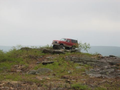 1990 Ford Bronco - A red SUV navigates a rocky incline under an overcast sky.