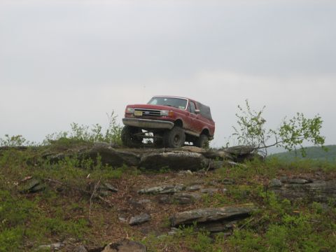 1990 Ford Bronco - Red 1990 Ford Bronco navigating a challenging rocky trail under a cloudy sky.