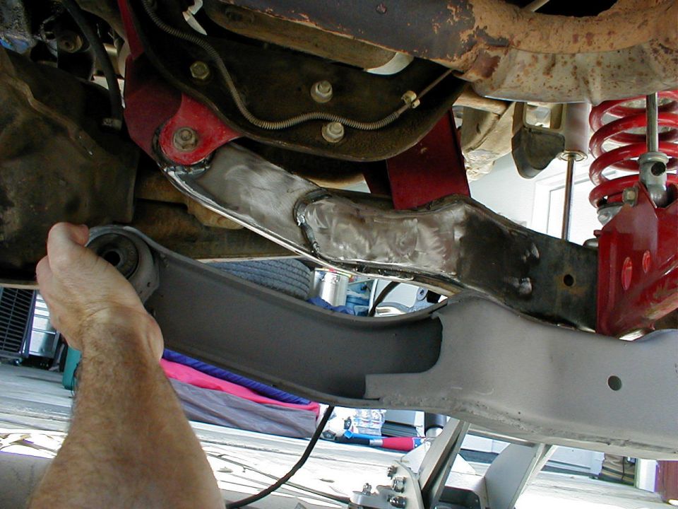 Close-up of a vehicle's front suspension with a hand holding a control arm. Welds are visible on a metal component