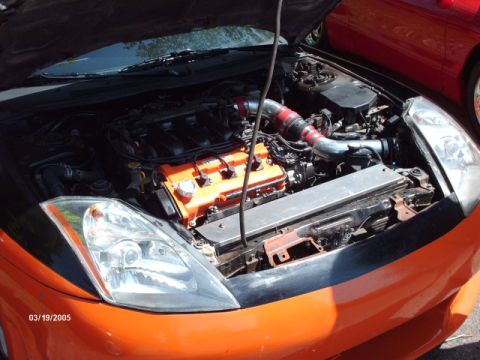 1995 Ford Probe - The engine bay of a car is open, revealing an orange-accented engine and custom intake piping. A red car is partially visible on the right.