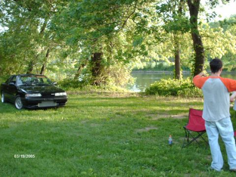 1995 Ford Probe - A black coupe with an aftermarket intercooler is parked on a grassy area next to a river, with a person standing nearby.
