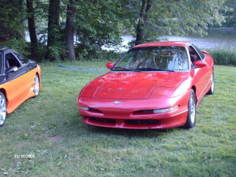 1995 Ford Probe - A red 1995 Ford Probe is parked on a grassy area near a body of water, with another vehicle visible to its left.