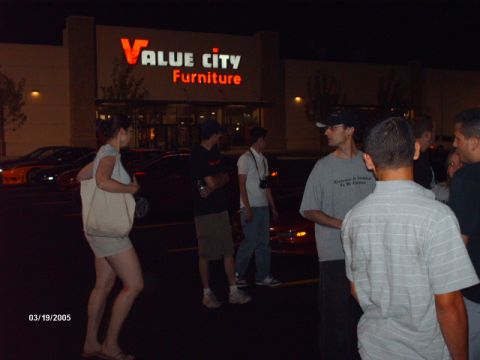 1995 Ford Probe - An orange sports car is visible at night in front of a Value City Furniture store, with several people standing nearby.