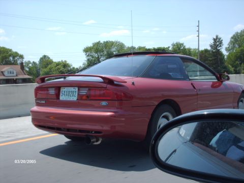 1995 Ford Probe - A red 1995 Ford Probe is seen from the rear, driving on a highway. Another vehicle's side mirror is visible in the foreground.