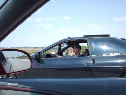1995 Ford Probe - A dark coupe with a driver is seen from another vehicle on a highway, with a sticker on the rear window.