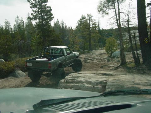1986 Ford Bronco - A heavily modified green pickup truck tackles a challenging rocky trail in a wooded area.