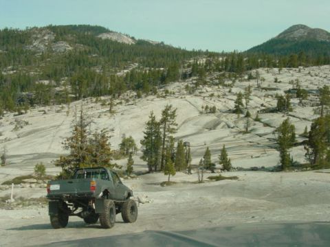 1986 Ford Bronco - A modified pickup truck is parked on a dirt road in a rocky, mountainous area with scattered trees.