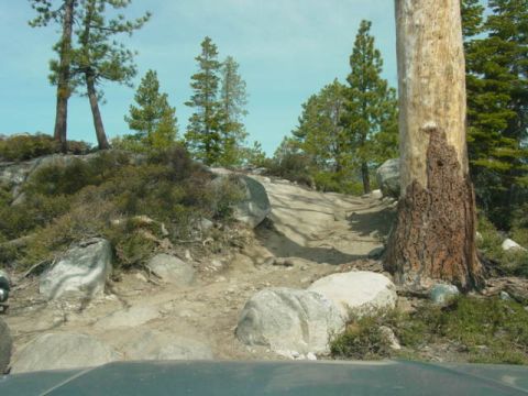 1986 Ford Bronco - View from inside a vehicle navigating a challenging, rocky off-road trail surrounded by trees and large rocks.