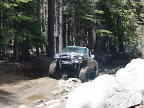 1986 Ford Bronco - A heavily modified pickup truck navigates a challenging off-road trail through a forest.