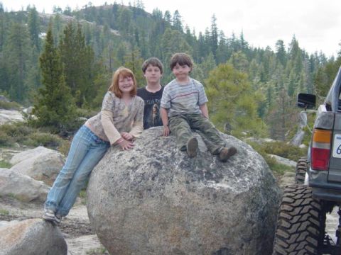 1986 Ford Bronco - Kids take a break on a large rock during an off-road excursion, with a vehicle parked close by.