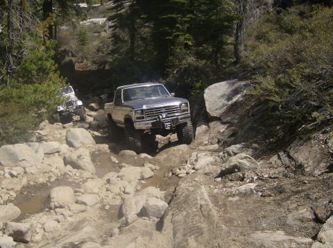 1986 Ford Bronco - Two off-road vehicles navigate a rocky, wooded trail. The lead vehicle is a Ford Bronco.