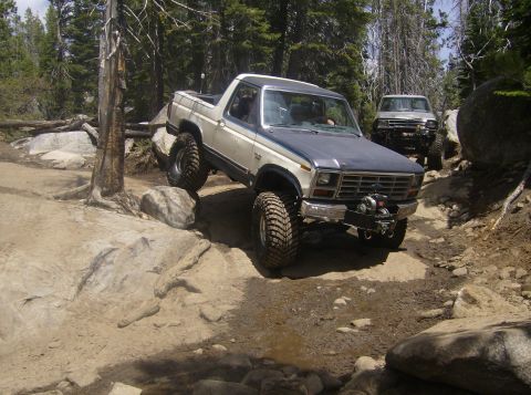 1986 Ford Bronco - Two off-road vehicles navigate a rocky trail through a wooded area.