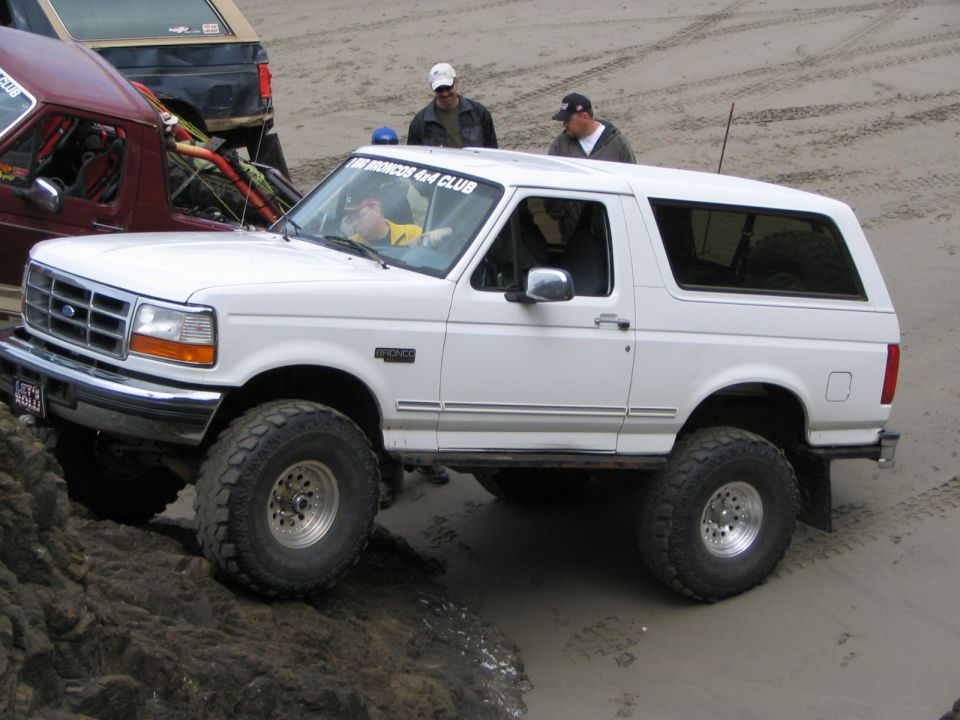 White 1990s Ford Bronco with lift and mud tires driving off a rock onto wet sand