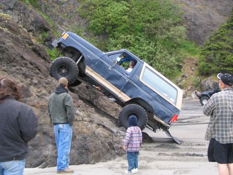 1983 Ford Bronco - A Ford Bronco navigates a challenging rocky terrain on a beach, with onlookers observing the off-road maneuver.