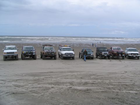 1983 Ford Bronco - A group of off-road vehicles are lined up on the sand near the ocean under a cloudy sky.
