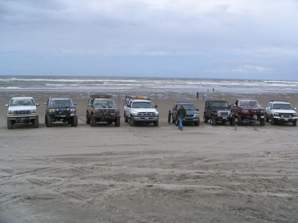 Eight lifted 4x4 trucks and Jeeps lined up nose-out on an overcast ocean beach