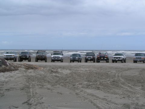 1983 Ford Bronco - Several off-road vehicles are parked in a line on a sandy beach with the ocean in the background.