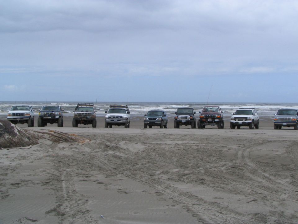 Row of nine lifted 4x4 trucks and Jeeps parked facing out on an overcast ocean beach