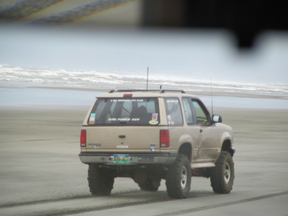 Lifted tan two-door Ford Explorer driving away on a wet ocean beach, rear view