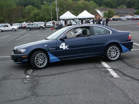 2001 Mercury Grand Marquis - A blue BMW coupe with racing decals and a driver in a helmet is parked on asphalt, with other cars and tents in the background.
