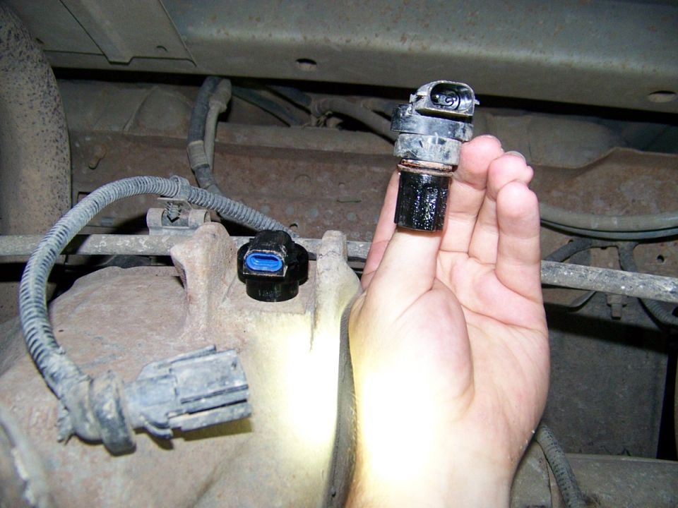 Hand holding a black speed sensor, with another sensor and wiring visible on a vehicle's undercarriage