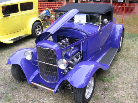 1989 Ford Mustang - A custom purple convertible hot rod is displayed with its hood open, revealing a Chevrolet engine, while a yellow car sits nearby.