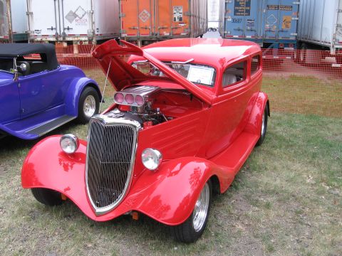 1989 Ford Mustang - A red hot rod sedan is displayed with its hood open, revealing a prominent supercharger and engine components.