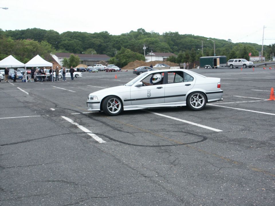 Silver BMW E36 sedan with racing number 5 on door, driver wearing helmet. Tires have left marks on asphalt