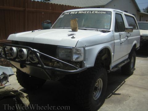 1990 Ford Bronco - white lifted Ford Bronco with a fabricated tube front bumper and round light pods parked by a wooden fence.