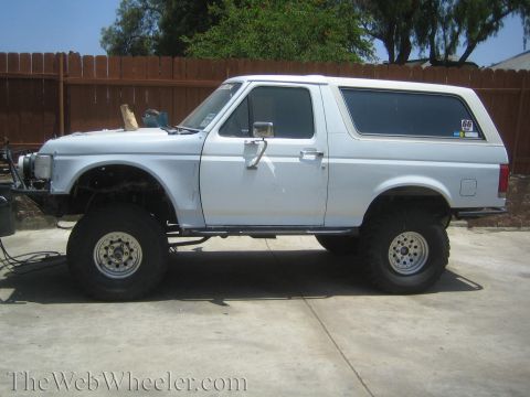 1990 Ford Bronco - Side profile of a white lifted Ford Bronco with hardtop and chrome off-road wheels parked on a driveway.