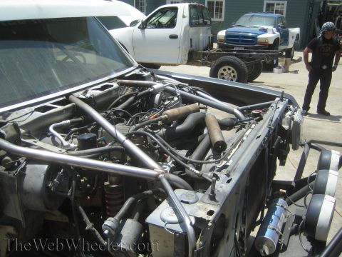 1990 Ford Bronco - tube-chassis race truck engine bay sits open as a man stands by, with a Dodge and white trucks in the background.
